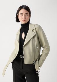 Jachetă de biker din piele verde deschis, cu buzunare cu fermoar, detalii metalice argintii și curea. Purtați-o peste un pulover negru cu guler înalt cu nervuri.