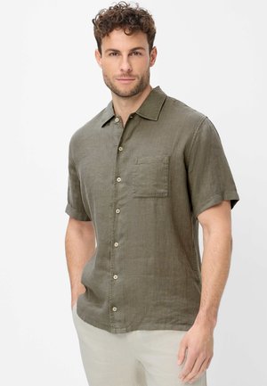 Mann mit lockigem Haar, der ein kurzärmeliges olivgrünes Hemd mit Knöpfen und helle Hosen trägt, steht mit einer Hand in der Tasche.