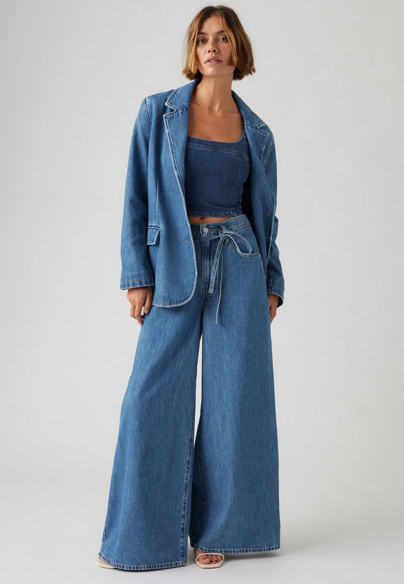 Tenue en denim comprenant une veste bleue ample et un jeans bleu à jambes larges. Le haut est un crop top en denim ajusté avec une finition texturée.