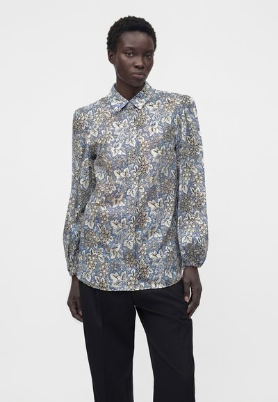 Femme portant un chemisier bleu à fleurs à manches longues boutonné et un pantalon noir, debout devant un fond blanc uni.