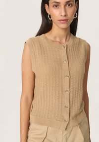 Beige ärmlös stickad cardigan med ett strukturerat mönster, rund halsringning och femknapps-stängning fram. Accentuerad med en ribbad nederkant.