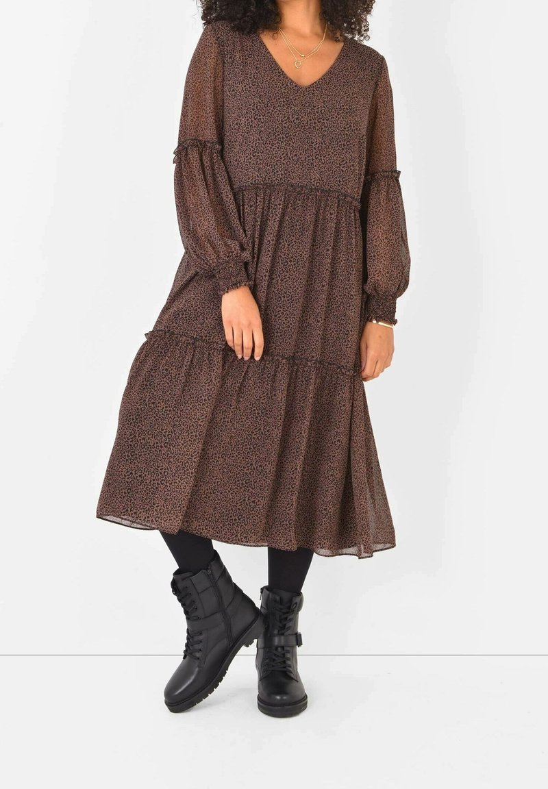 Mujer con un vestido midi marrón de estampado, con mangas largas transparentes, medias negras y botas de combate negras con cordones, de pie con las piernas cruzadas.