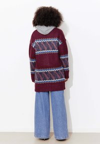 Cardigan en tricot bordeaux avec un motif géométrique en bleu, violet et crème. Il comprend une capuche grise ainsi que des poignets et une bordure côtelés. Associé à un jean à jambes larges.