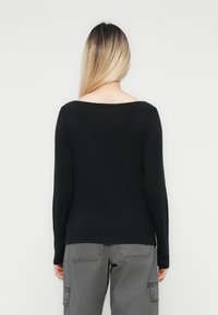 Persoon met lang blond haar, gekleed in een zwart shirt met lange mouwen en grijze cargobroek, staand met de rug naar de camera tegen een effen achtergrond.