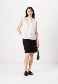 Ärmellose weiße Bluse aus strukturiertem Stoff mit Stehkragen und Knopfleiste; kombiniert mit schwarzen Shorts und schwarzen Mokassins. Kleine schwarze Handtasche.