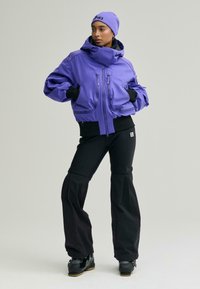 Lila pufi dzseki cipzáras zsebekkel, fekete bordázott mandzsettákkal és hozzáillő fekete nadrággal, letisztult dizájnnal és széles szárnyílásokkal.