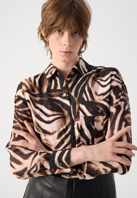 Camicia a maniche lunghe in tessuto leggero con stampa zebra marrone e nera. Presenta una chiusura con bottoni sul davanti e una tasca sul lato sinistro del petto.