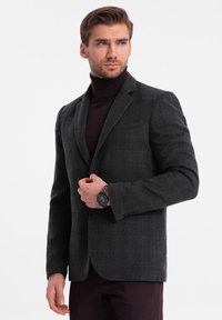 Grå hundtandsmönstrad blazer med slagkragar, skräddarsydd passform och två framfickor. Bärs över en mörk polotröja, tillsammans med mörka byxor.