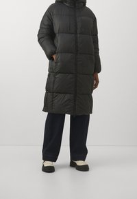 Sort polstret lang frakke med hætte, der har en frontlynlås, to sidelommer og en glat, blank overflade. Parret med løse mørke jeans.