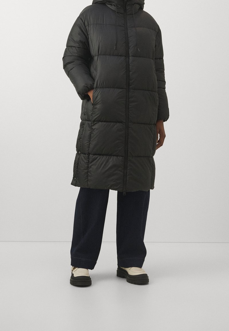 Sort polstret lang frakke med hætte, der har en frontlynlås, to sidelommer og en glat, blank overflade. Parret med løse mørke jeans.