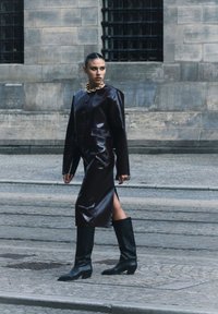 Veste en cuir marron courte avec de grands boutons, assortie à une jupe en cuir marron moulante avec une fente sur le côté. Bottes noires jusqu'au genou.