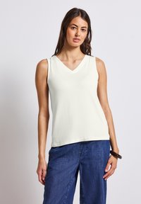 Top senza maniche, con scollo a V di colore crema chiaro. Tessuto morbido con una trama liscia. Abbinato a pantaloni larghi di colore blu scuro.