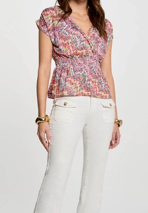 Femme portant un chemisier à manches courtes avec un motif en zigzag coloré, une taille froncée, et un pantalon blanc avec des poches avant à boutons.