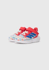 Une paire de baskets pour tout-petits avec des sangles rouges et des accents bleus, présentant une base blanche ornée de motifs abstraits rouges et bleus.