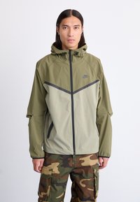 Letvægts, olivengrøn Nike-jakke med lynlås i front, hætte og kontrasterende mørkegrå detaljer. Kombineret med cargo-bukser i camouflagemønster.