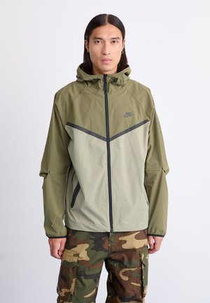 Lichte, olijfgroene Nike jas met een rits aan de voorkant, capuchon en contrasterende donkergrijze accenten. Gecombineerd met camouflagepatroon cargobroek.