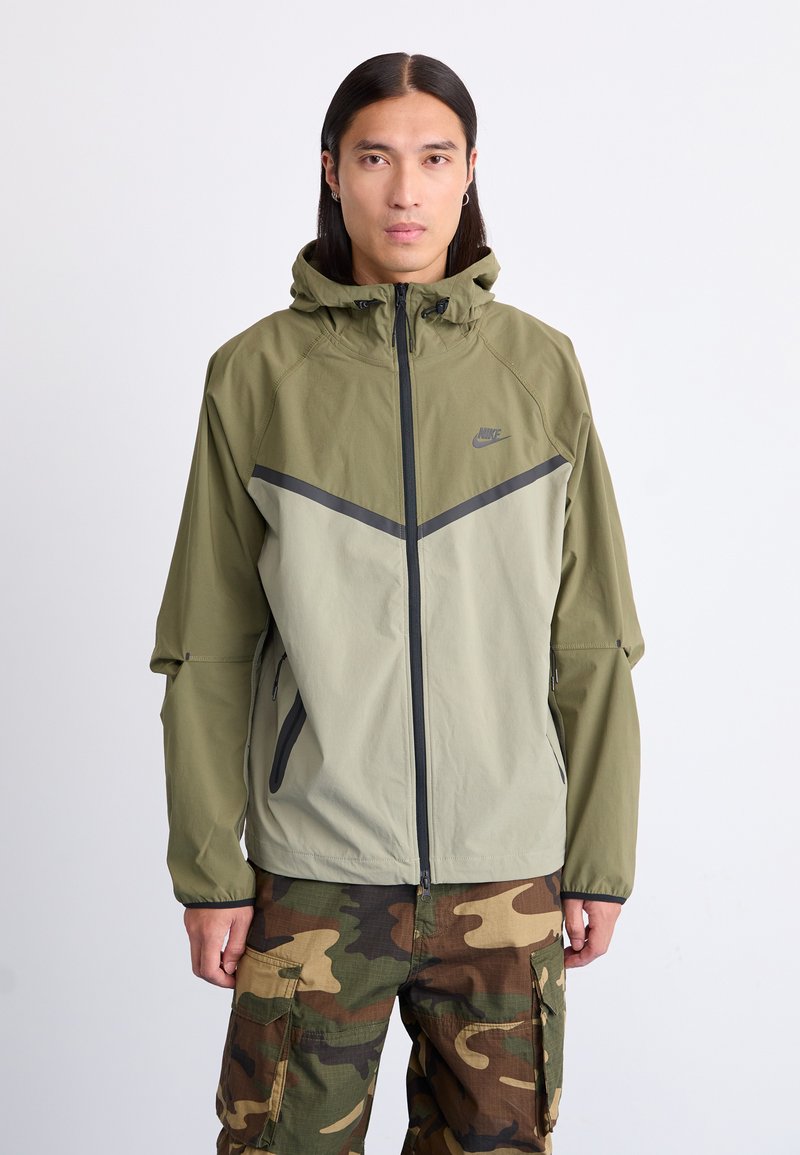 Letvægts, olivengrøn Nike-jakke med lynlås i front, hætte og kontrasterende mørkegrå detaljer. Kombineret med cargo-bukser i camouflagemønster.