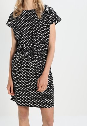 Nainen pukeutuneena mustaan, polvipituiseen mekkoon, jossa on lyhyet hihat ja valkoinen pieni kukkakuvio, vyötäröllä kiristysnauhalla sidottu.