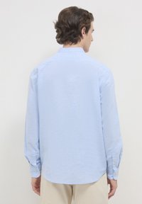 Uomo con capelli ricci castani che indossa una camicia azzurro chiaro con maniche lunghe e pantaloni beige, rivolto di spalle contro uno sfondo neutro.