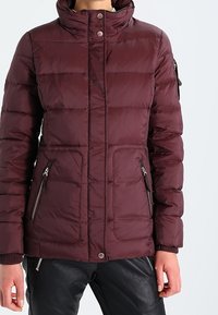 Burgundy pufferjakke med høj krave, lynlåslommer i siderne, knaplukning og ribbede manchetter, med quiltet design og blød tekstur.