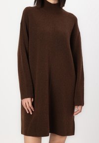 Hnědá pletená šaty s vysokým výstřihem, oversized střihem a dlouhými rukávy. Měkká textura a žebrované manžety umocňují design.