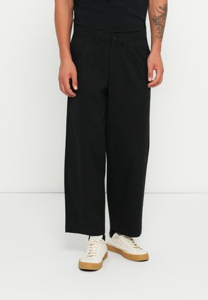Black wide-leg trousers made of cotton, featuring a button closure and side pockets. Paired with white sneakers with a tan rubber sole.