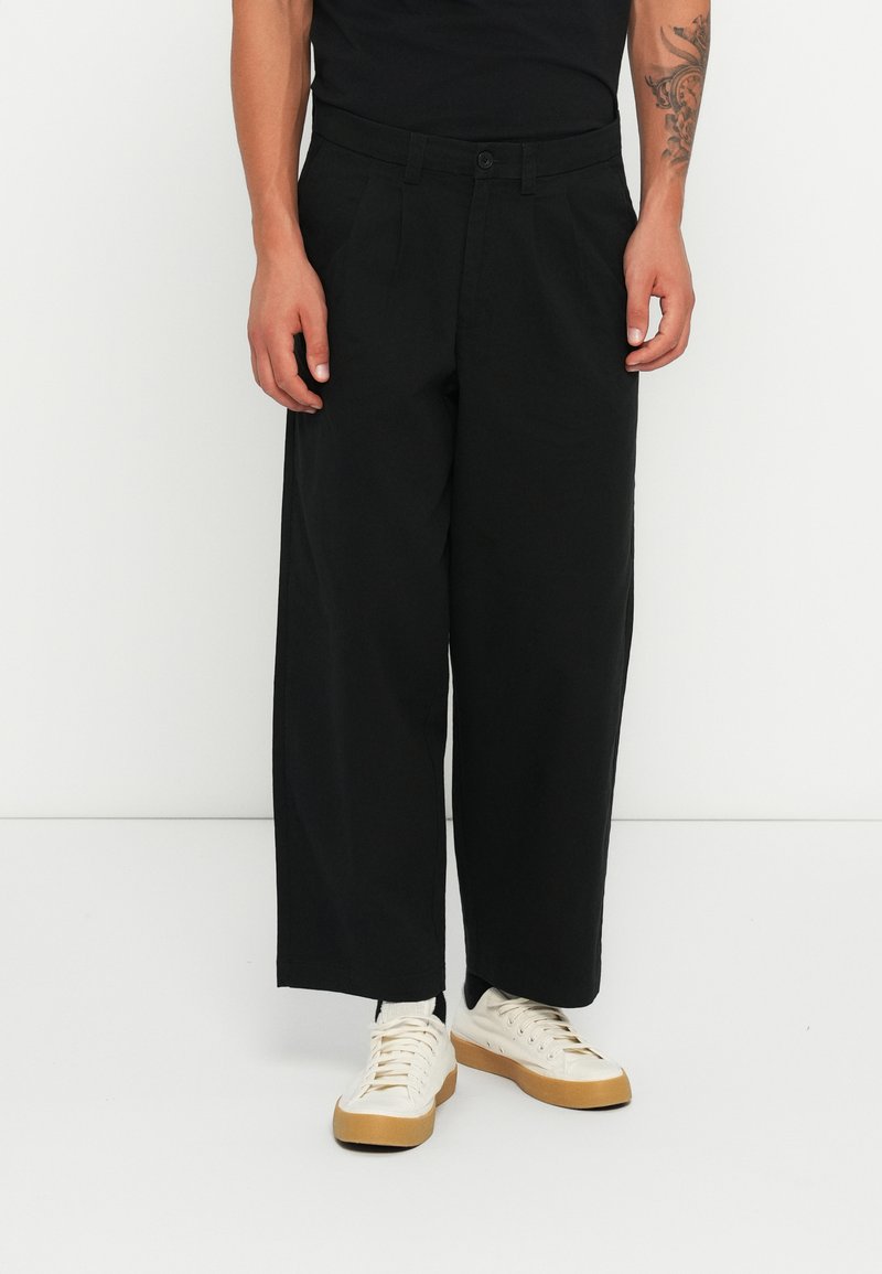 Black wide-leg trousers made of cotton, featuring a button closure and side pockets. Paired with white sneakers with a tan rubber sole.