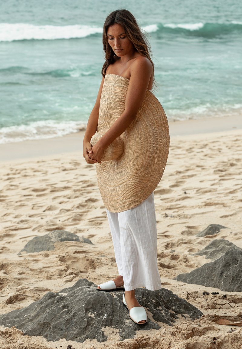 Chapeau à large bord en paille, pantalon en lin blanc cassé et sandales à bout ouvert sur une plage rocheuse. Vagues de l'océan en arrière-plan, plage de sable au premier plan.