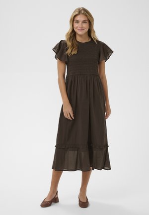 Žena v tmavohnedom midi šatách s krátkymi volánovými rukávmi, naškrobeným živôtikom, volánikovým okrajom a nízkymi blokovými topánkami z tkaniny, stojaca pred bielym pozadím.