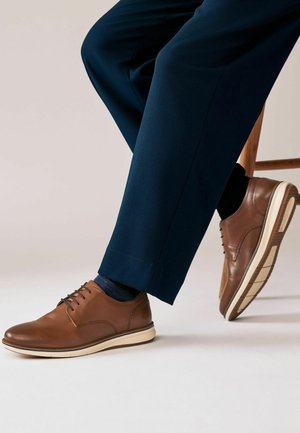 Chaussures en cuir marron à lacets avec un bout arrondi, semelle de couleur crème et détails de coutures subtils, associées à un pantalon bleu marine et des chaussettes sombres.
