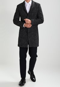 Svartvit kappa med fiskbensmönster, skräddarsydd passform, stängning med en knapp, kombinerad med mörka byxor och svarta skor. Slät textur.