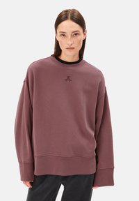 Frau mit glattem braunem Haar tr�ägt einen übergroßen mauvefarbenen Pullover mit kleinem gesticktem Logo und steht vor einem schlichten Hintergrund.