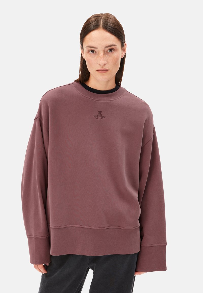 Frau mit glattem braunem Haar trägt einen übergroßen mauvefarbenen Pullover mit kleinem gesticktem Logo und steht vor einem schlichten Hintergrund.