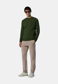 Maglione verde lavorato a maglia, pantaloni beige e sneakers bianche. Il maglione presenta polsini e orlo a coste. La modella indossa occhiali da sole scuri.
