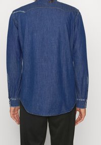 Camicia di jeans blu con maniche lunghe, orlo curvo e cuciture in contrasto. Polsini con bottoni e dettaglio sul giromanica posteriore.