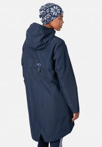 Marineblauwe waterdichte jas met capuchon, met een relaxte pasvorm, verstelbare taille en versterkte stiksels. Gecombineerd met een gestreepte blauwe beanie.