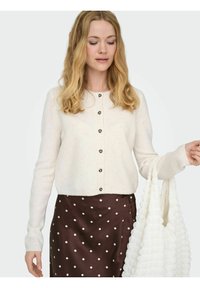 Cardigan crema chiaro con bottoni neri, caratterizzato da un collo rotondo e maniche lunghe, abbinato a una gonna marrone con pois e una borsa bianca texturizzata.