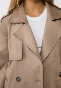 Beige semišový trenčkot s širokými klopami, dvouřadým zapínáním a tmavými knoflíky. Nosený přes bílou žebrovanou košili.