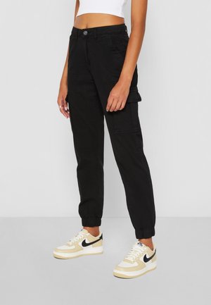 Femme portant un pantalon cargo jogging noir avec des poignets élastiques et des baskets Nike beige, debout devant un fond uni gris clair.