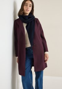 Manteau maroon à l'avant ouvert, fermeture à un bouton unique, porté sur un pull léger et un jean bleu. Écharpe sombre enroulée autour du cou.
