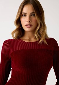 Maglione bordeaux a coste con maniche lunghe; il design testurizzato presenta costole orizzontali e verticali, un colletto arrotondato e una finitura morbida.
