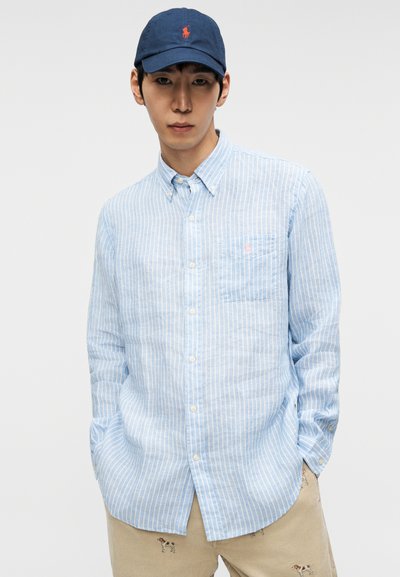 Light blue striped button-up shirt with a chest pocket and a small pink logo. Paired with a navy baseball cap. Casual, relaxed fit.