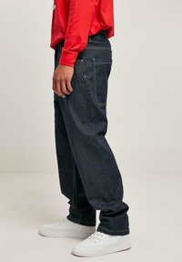 Jeans in denim blu scuro con una vestibilità rilassata, cuciture visibili, tasche laterali e abbinati a sneakers bianche. È visibile una maglietta a maniche lunghe rossa.