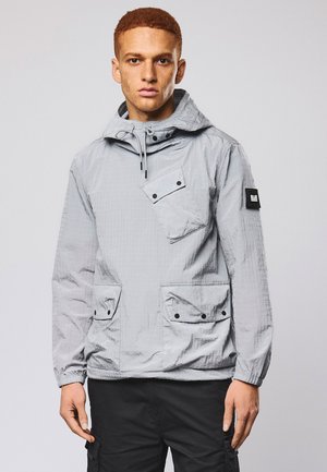 Mand med kort krøllet hår iført en lysegrå hættejakke med tre knaplommer og sorte bukser foran en ensfarvet baggrund.