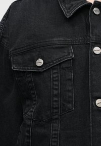 Casaco de ganga preto com gola, dois bolsos no peito, ferragens prateadas e detalhes de costura visíveis. Tecido texturado.