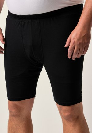 Uomo in boxer neri fino al ginocchio con cintura elastica e una camicia bianca, in piedi con le mani rilassate ai lati.