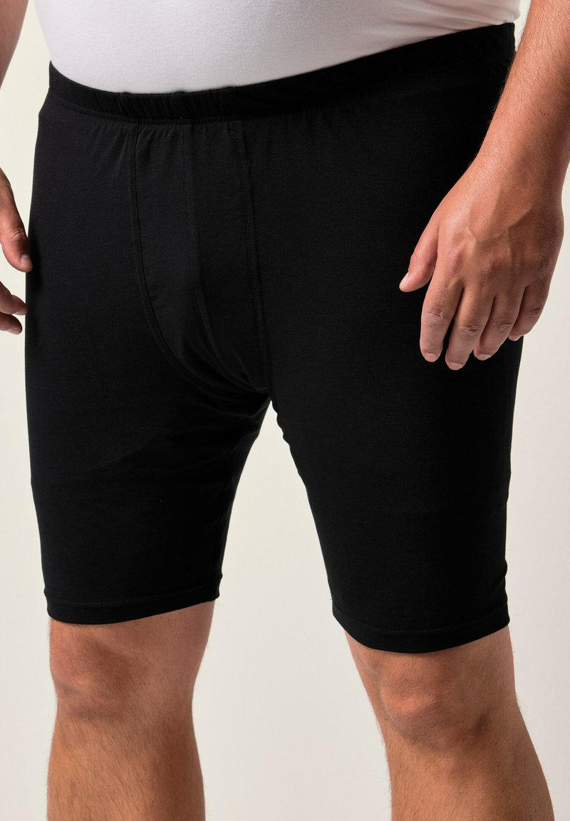 Uomo in boxer neri fino al ginocchio con cintura elastica e una camicia bianca, in piedi con le mani rilassate ai lati.