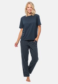 Donna sorridente in piedi a piedi nudi, che indossa una maglietta blu navy a maniche corte e pantaloni blu navy con strisce verticali bianche, con una mano in tasca.