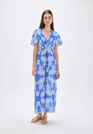 Mujer de pie, con un mono azul con un llamativo estampado floral blanco, mangas cortas, pendientes dorados y sandalias doradas sobre un fondo liso.