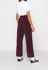 Pantalon en velours côtelé bordeaux à jambes larges, taille haute et poches arrière, associé à un t-shirt blanc et des baskets noires.